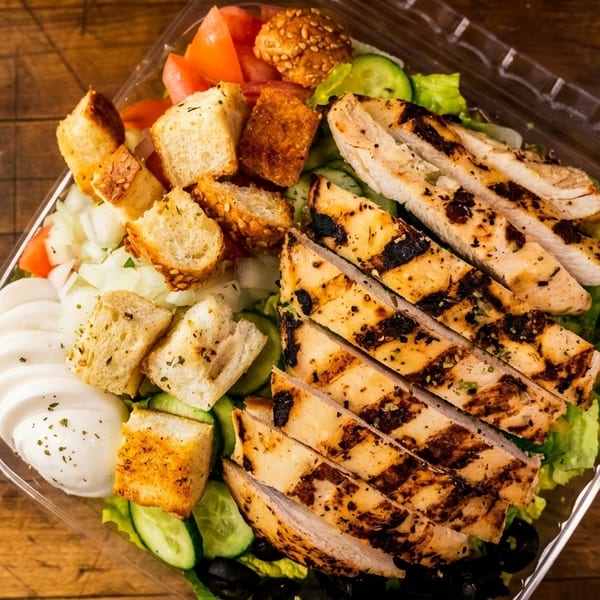 Grilled chicken salad with croutons, tomatoes, cucumbers, and mozzarella in a plastic container.