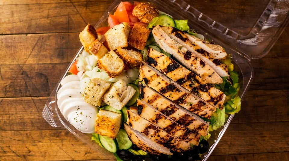Grilled chicken salad with croutons, tomatoes, cucumbers, and mozzarella in a plastic container.
