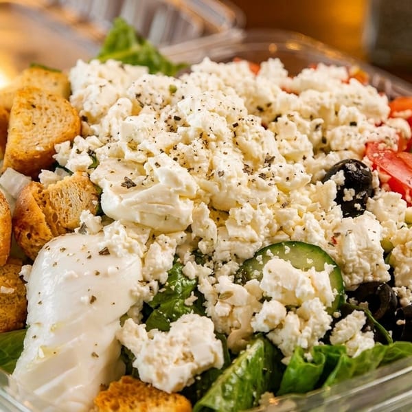 A close-up of a vibrant Greek salad in a clear plastic container, topped with feta cheese.