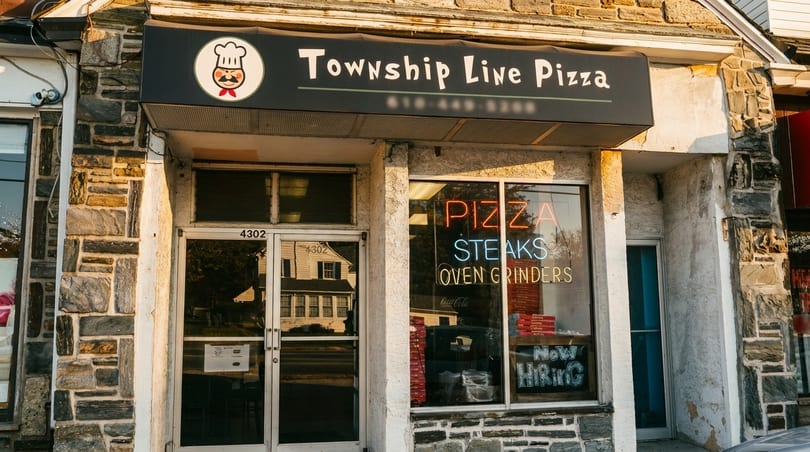 Exterior of Township Line Pizza, with a black awning and neon "PIZZA STEAKS" sign in the window.