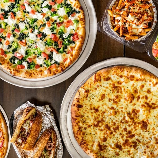 An overhead view of a feast of pizzas, subs, loaded fries, and a grilled chicken salad.