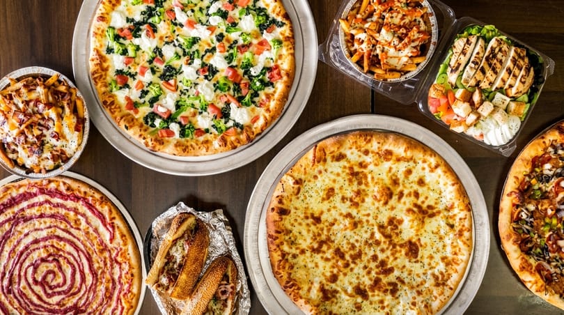 An overhead view of a feast of pizzas, subs, loaded fries, and a grilled chicken salad.