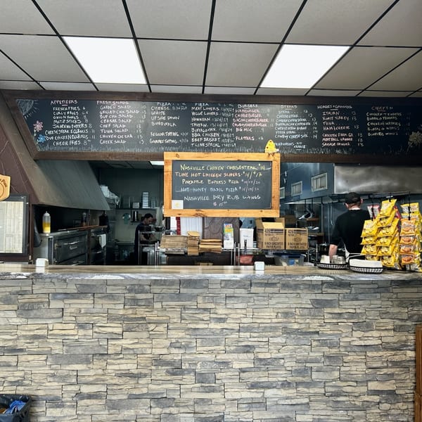 A restaurant interior with a stone counter, a large menu chalkboard, and kitchen staff working.