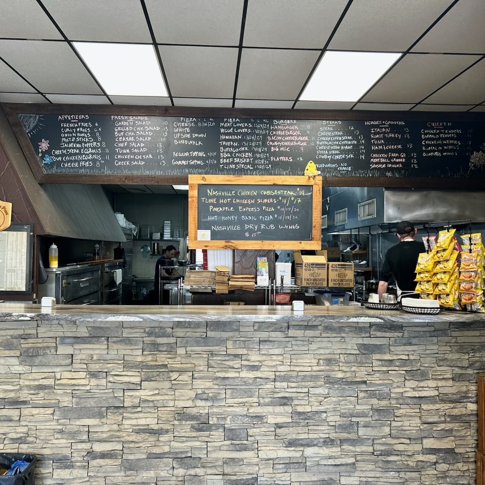 A restaurant interior with a stone counter, a large menu chalkboard, and kitchen staff working.
