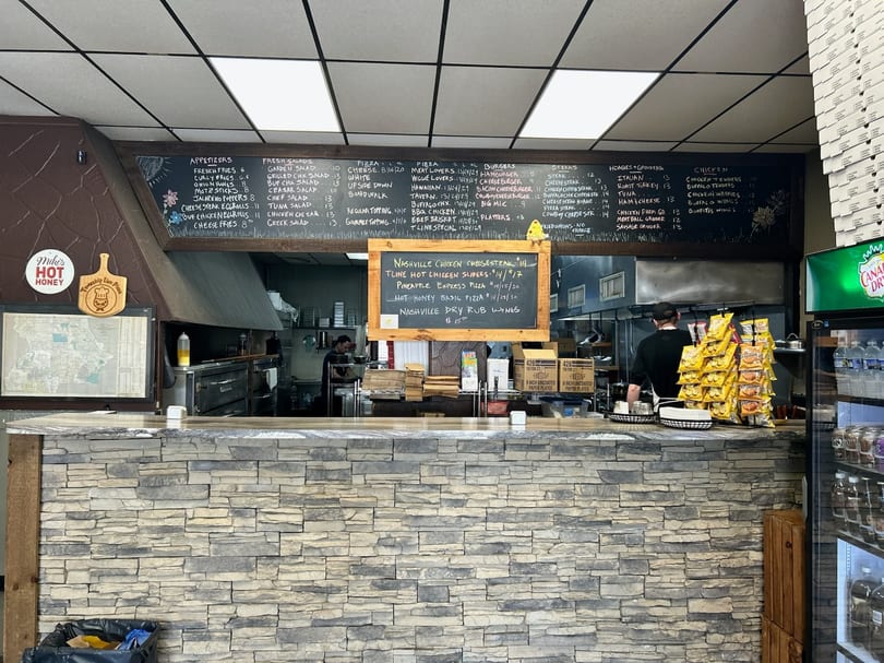 A restaurant interior with a stone counter, a large menu chalkboard, and kitchen staff working.