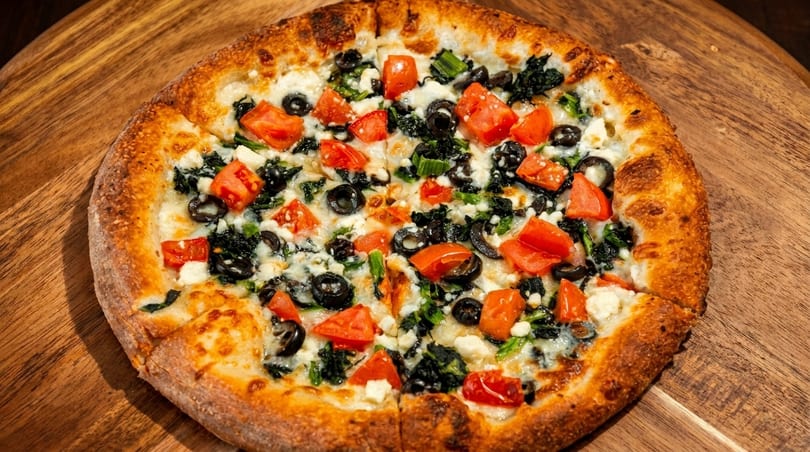 A whole pizza with spinach, black olives, diced tomatoes, and feta cheese on a wooden board.
